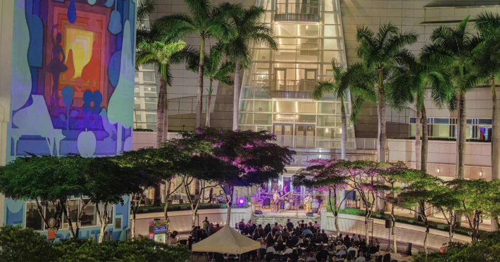 Una actuación cultural hispana al aire libre en el Adrienne Arsht Center, con músicos actuando en el escenario mientras el público observa desde la plaza del centro, bajo las palmeras.