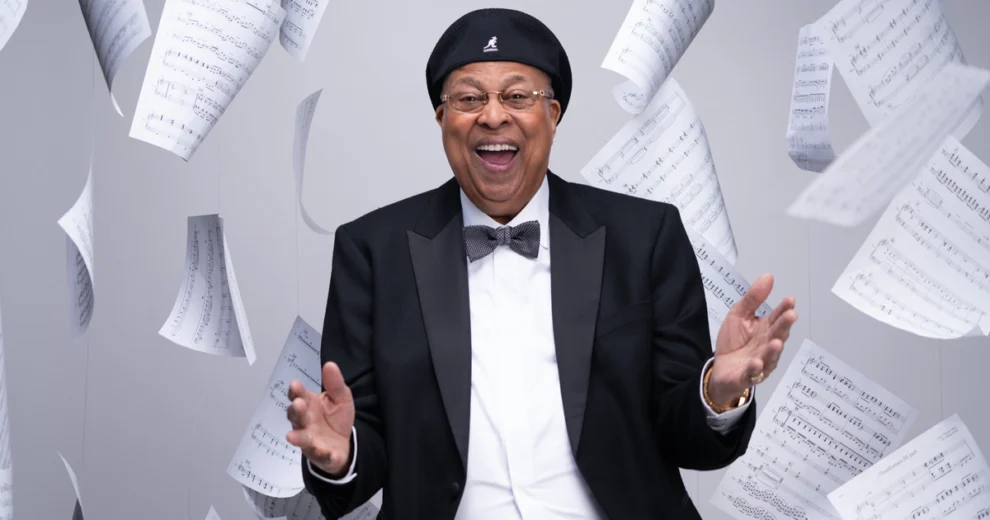 Un músico negro sonriente, vestido con esmoquin, pajarita, gafas y gorra negra, posa entre hojas de partituras flotantes como parte de la programación hispana del Adrienne Arsht Center.