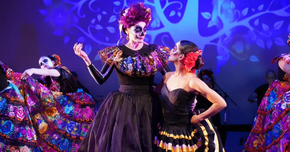 Una colorida actuación cultural hispana en el Adrienne Arsht Center, con bailarines que lucen elaborados trajes y maquillaje inspirado en el Día de los Muertos sobre un escenario iluminado de azul.