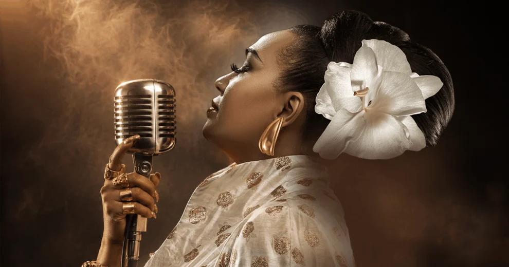 Grammy and Latin Grammy-winning vocalist Aymée Nuviola sings into a vintage microphone, wearing a white patterned outfit, gold jewelry, and a large white flower in her hair.