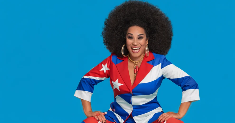 Grammy and Latin Grammy-winning vocalist Aymée Nuviola smiles at the camera while wearing a colorful suit inspired by the Cuban flag, with a bright blue background.