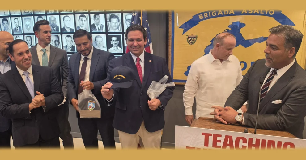 Several people gather inside the same memorial setting. A man in a dark suit holds a commemorative cap and gift bag, while others stand nearby. Behind them are portraits of individuals and a large “Brigada Asalto” display. A podium sign in the foreground partially reads “Teaching the Dangers…”