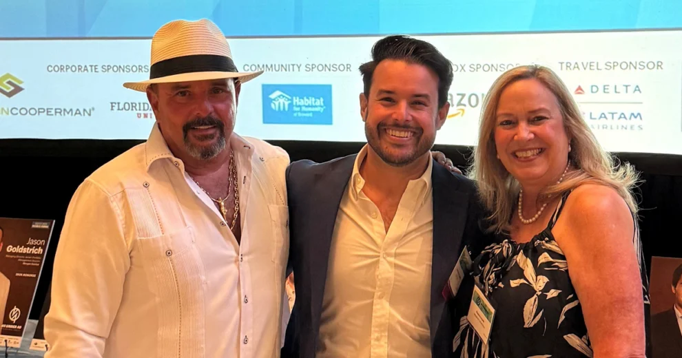 Branden Lopez posa con otros dos asistentes en la ceremonia de premios "40 Under 40", con los patrocinadores del evento mostrados en una pantalla de presentación al fondo.