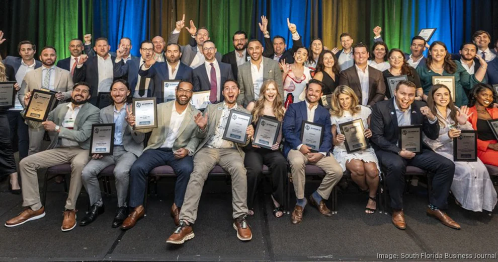 Branden Lopez and other 40 Under 40 Awards honorees pose onstage while holding plaques and certificates. Colorful blue and green curtains are visible behind the group.
