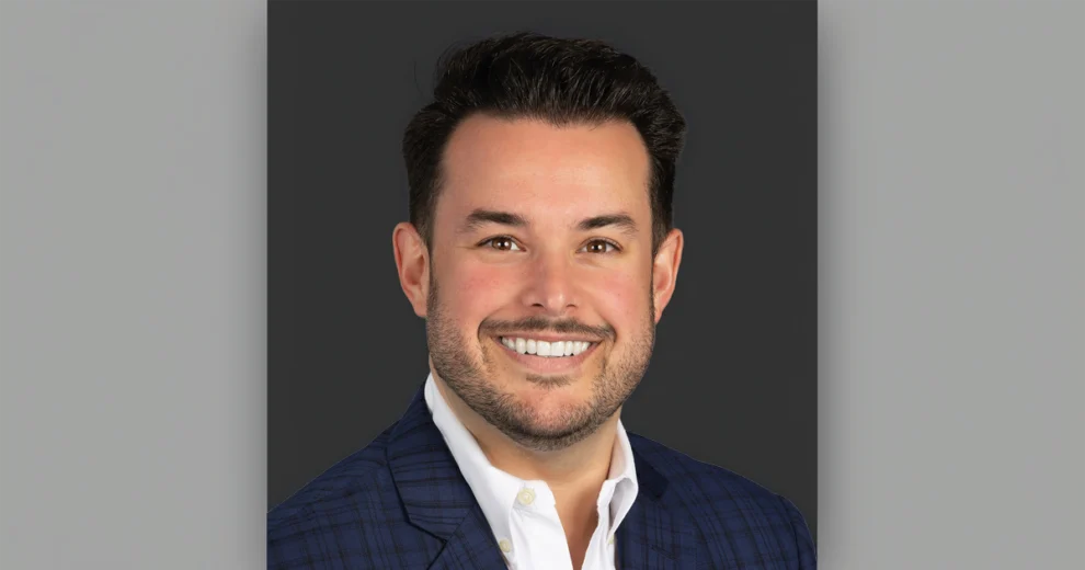 Professional headshot of Branden Lopez smiling and wearing a navy plaid suit jacket and white dress shirt against a dark gray background.