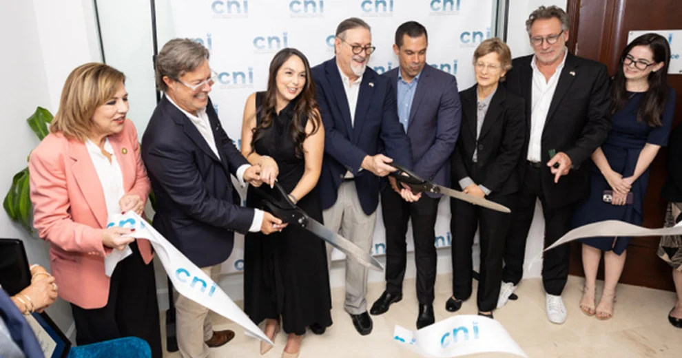 Un grupo de adultos participa en una ceremonia de corte de cinta frente a un telón de fondo con la marca CNI, sosteniendo unas tijeras ceremoniales de gran tamaño.