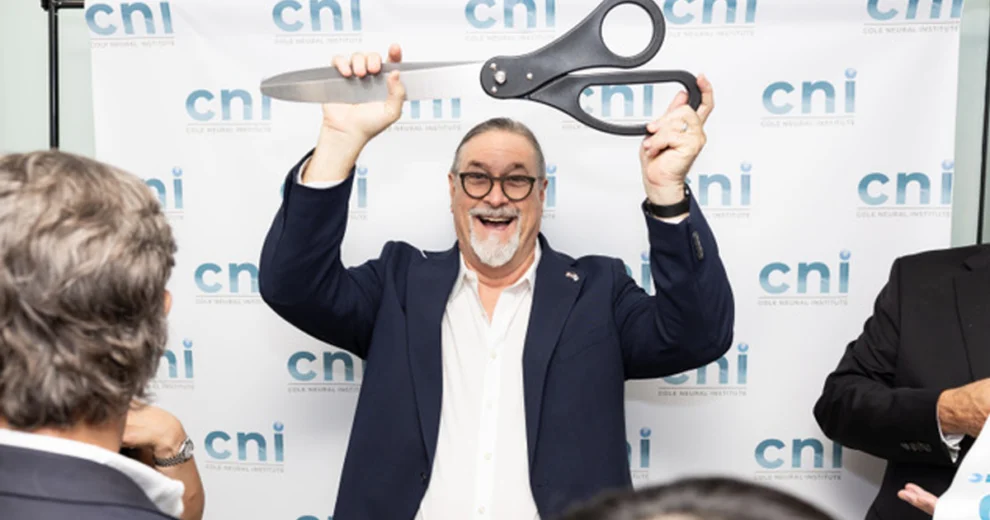 Un hombre sonríe mientras sostiene unas tijeras ceremoniales de gran tamaño sobre su cabeza, frente a un telón de fondo con la marca CNI.