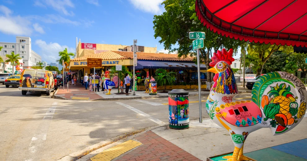 Colorful street scene in Miami’s Little Havana along Calle Ocho, featuring Cuban restaurants, colorful storefronts, pedestrians, palm trees, street signs, and brightly painted rooster sculptures.