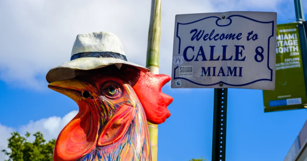 Primer plano de una escultura de gallo pintada con colores vivos y con sombrero, situada junto a una señal de calle que dice "Welcome to CALLE 8 MIAMI" en Little Havana, con el cielo azul y vegetación tropical de fondo.