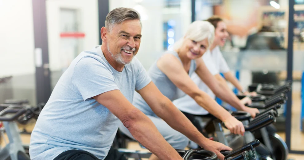 Un grupo de adultos mayores haciendo ejercicio en bicicletas estáticas en un gimnasio, sonriendo y participando en una sesión de entrenamiento físico.