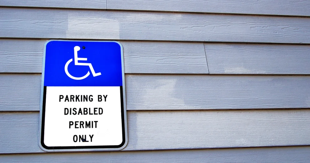 Una señal de estacionamiento accesible de color azul y blanco, fijada en un edificio, dice "Estacionamiento exclusivo para vehículos con permiso de discapacidad" e incluye el símbolo internacional de accesibilidad para sillas de ruedas.
