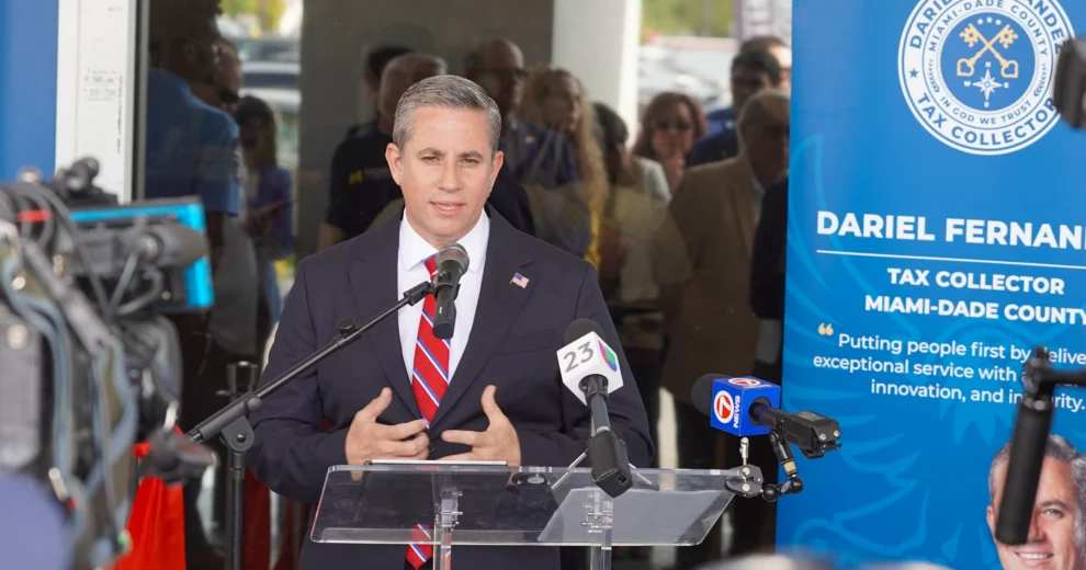 El recaudador de impuestos del condado de Miami-Dade, Dariel Fernandez, habla desde un podio durante una conferencia de prensa, rodeado de cámaras y micrófonos de los medios de comunicación. Al fondo se puede ver una pancarta de la oficina del recaudador de impuestos del condado de Miami-Dade.