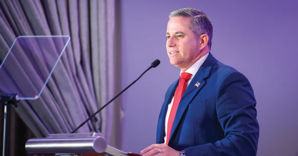 Miami-Dade County Tax Collector Dariel Fernandez speaks at a podium during an indoor event, wearing a blue suit and red tie. A teleprompter and microphone are visible beside him against a purple-lit backdrop.