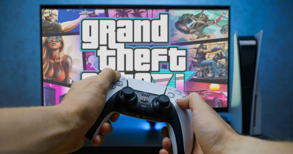 A person holding a PlayStation controller while playing Grand Theft Auto on a television. The screen displays the GTA VI logo and game artwork, with a PlayStation console visible beside the TV.