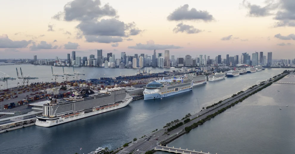 Aerial view of PortMiami with several large cruise ships docked alongside the container terminal, with the Miami skyline visible across Biscayne Bay.