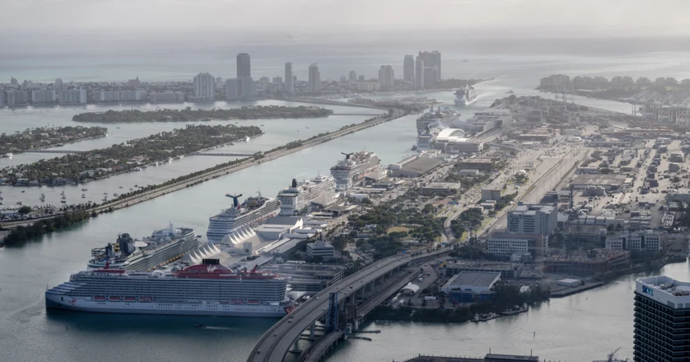Aerial view of PortMiami showing multiple cruise ships docked at terminals, cargo containers and cranes, the MacArthur Causeway, Biscayne Bay, and the Miami Beach skyline in the distance.