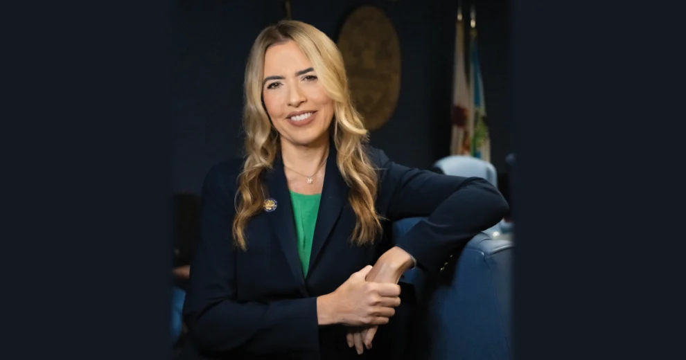 La comisionada del condado de Miami-Dade, Raquel Regalado, sonríe mientras está sentada en una sala de reuniones gubernamental, vistiendo una chaqueta azul marino sobre una blusa verde. Al fondo se aprecian un sello oficial y varias banderas.