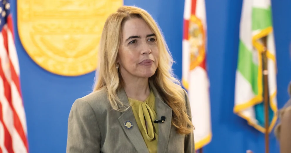Raquel Regalado Calls on Miami-Dade Employers to Turn Inclusion into Jobs 1 Miami-Dade County Commissioner Raquel Regalado stands at a government event wearing a gray blazer and olive-green blouse, with U.S., Florida, and Miami-Dade County flags and a large official seal visible in the background.