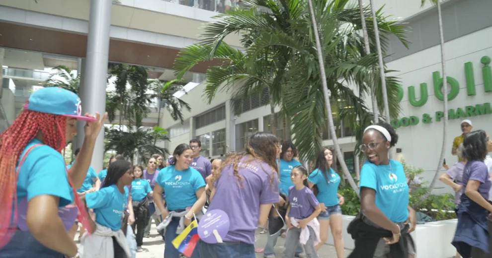 Members of Vocal Youth Miami, wearing turquoise and purple shirts, walk through the atrium at River Landing Shops & Residences during a group performance.