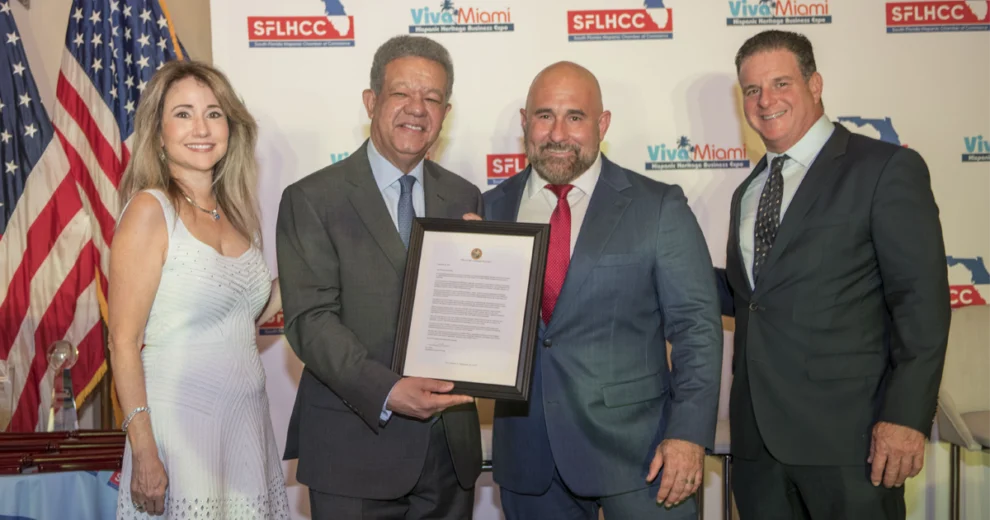 Four people pose at a South Florida Hispanic Chamber of Commerce event, with one man holding a framed proclamation. U.S. and Florida flags and SFLHCC and Viva Miami logos are displayed in the background.