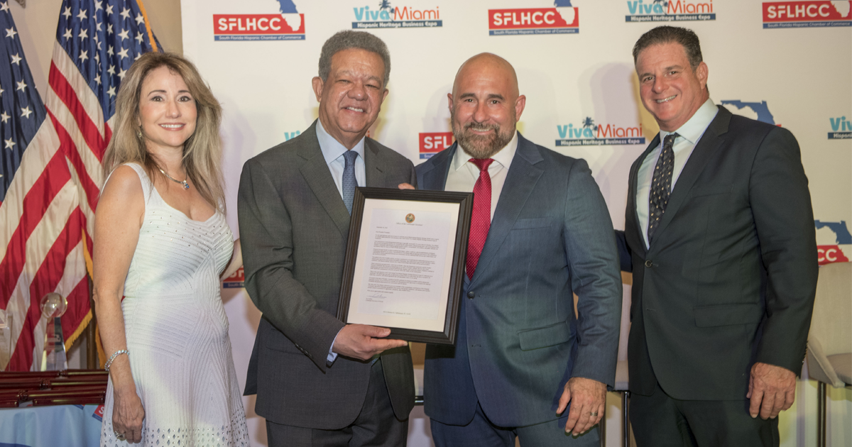 Cuatro personas posan en un evento de la Cámara de Comercio Hispana del Sur de Florida; uno de los hombres sostiene una proclamación enmarcada. De fondo se exhiben las banderas de Estados Unidos y Florida, así como los logotipos de la SFLHCC y Viva Miami.