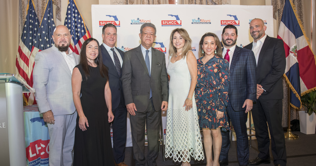 Eight South Florida Hispanic Chamber of Commerce event attendees pose together in front of an SFLHCC and Viva Miami backdrop, with U.S. and Florida flags displayed on both sides.