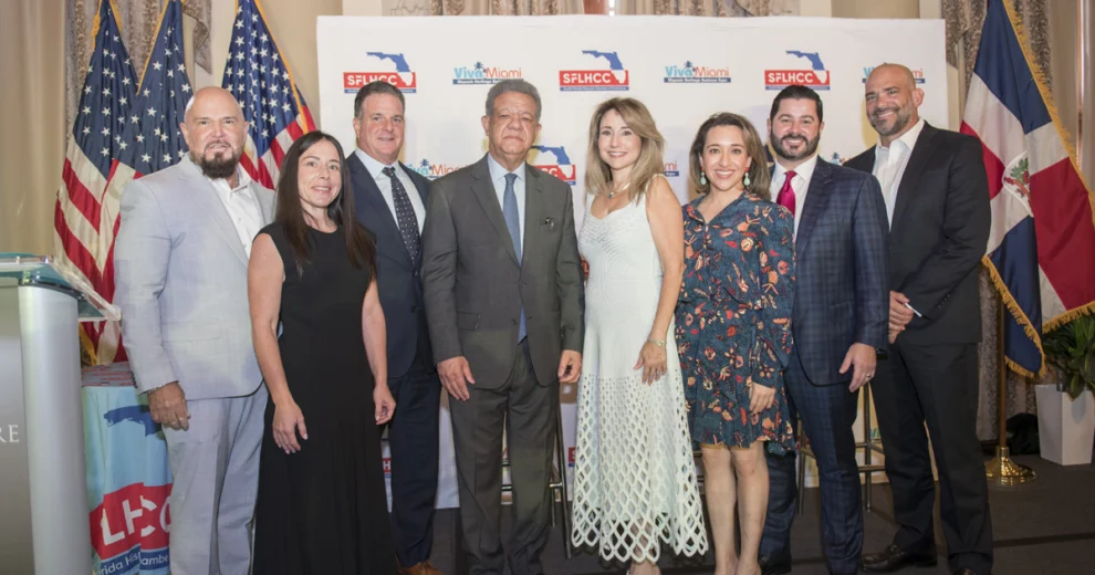 Ocho asistentes a un evento de la Cámara de Comercio Hispana del Sur de Florida posan juntos frente a un telón de fondo de la SFLHCC y Viva Miami, con las banderas de Estados Unidos y de Florida exhibidas a ambos lados.