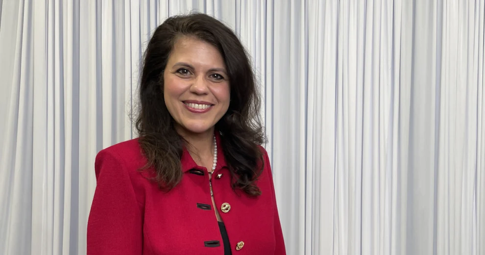 Senator Ana Maria Rodriguez smiles at the camera wearing a bright red jacket with gold buttons and a pearl necklace, standing in front of white curtains.