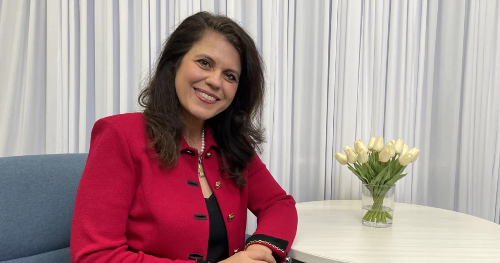 Senator Ana Maria Rodriguez sits at a white table wearing a bright red jacket and pearl necklace, smiling at the camera. A vase of white tulips sits on the table beside her, with white curtains and a blue upholstered chair in the background.
