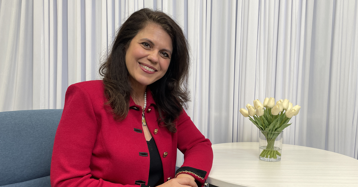 Senator Ana Maria Rodriguez sits at a white table wearing a bright red jacket and pearl necklace, smiling at the camera. A vase of white tulips sits on the table beside her, with white curtains and a blue upholstered chair in the background.
