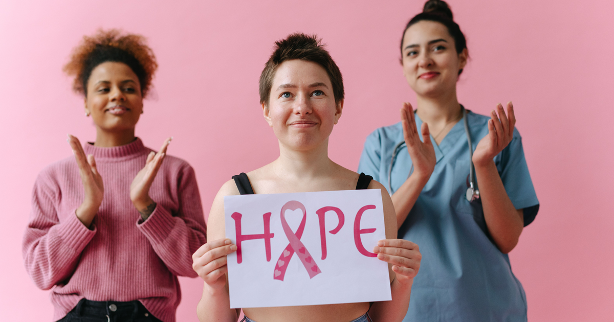 Three women stand against a pink background; the woman in front holds a sign reading &ldquo;HOPE&rdquo; with a pink breast cancer awareness ribbon, while the two women behind her applaud.