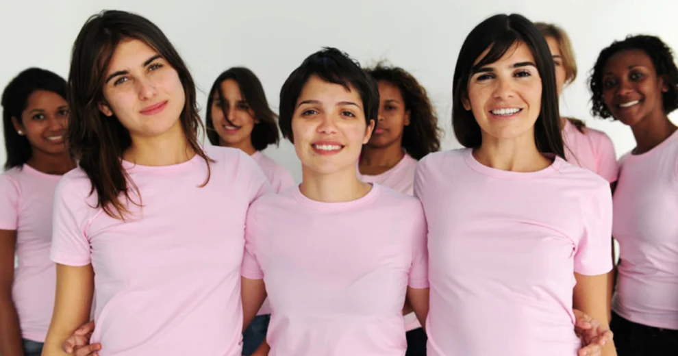 A diverse group of women wearing pink shirts stand together and smile at the camera, symbolizing breast health and breast cancer awareness.