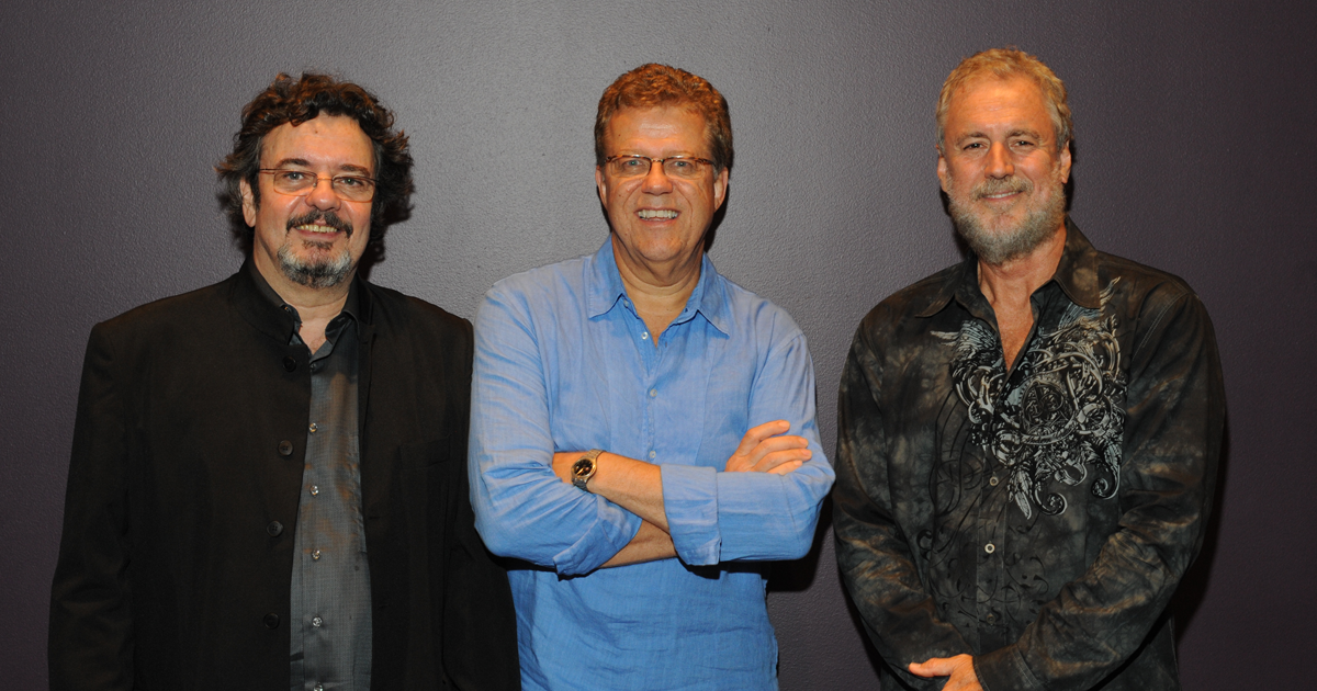 Trio da Paz poses for a group photo, with three male musicians standing side by side against a dark gray background at the Adrienne Arsht Center for the Performing Arts in Miami.