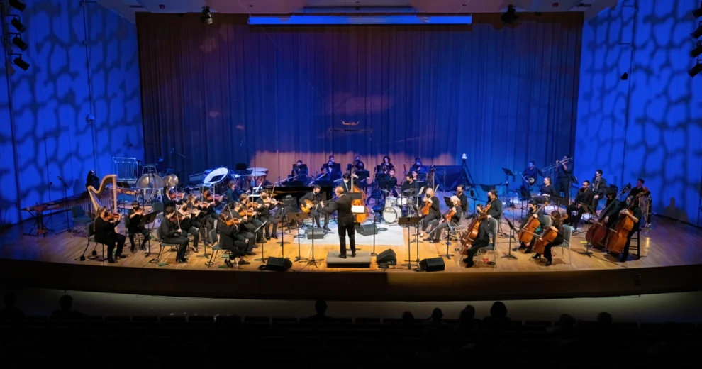 The Henry Mancini Institute Orchestra performs onstage at the Adrienne Arsht Center for the Performing Arts in Miami. Musicians play strings, brass, percussion, and other instruments under a conductor’s direction in a concert hall illuminated with blue stage lighting playing for the Jobim tribute.