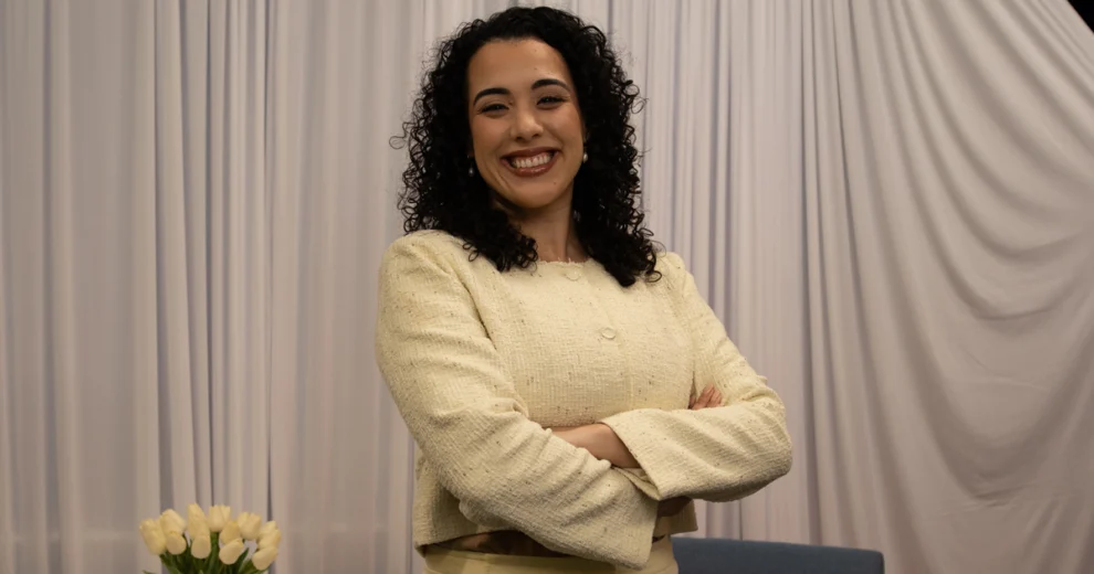 Florida Senator Alexis Calatayud poses with her arms crossed, wearing a cream-colored textured jacket and matching light-colored trousers with pearl earrings. White tulips sit in the lower-left corner, with a blue chair and light-colored draped curtains in the background.