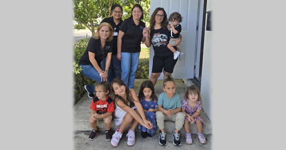 A group of adults and young children pose together outside a stable housing home, with several adults standing behind five children seated on the front steps.