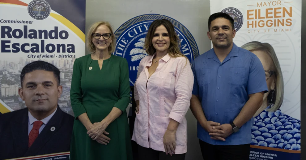 Calle Ocho News Editor-in-Chief Rosi Rosell, wearing a light pink blouse, poses with a woman in a green dress and glasses and a man in a blue shirt in front of City of Miami banners featuring Commissioner Rolando Escalona and Mayor Eileen Higgins.