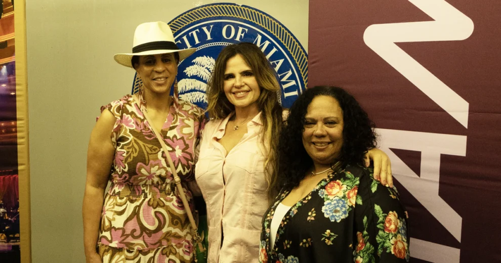 Rosi Rosell, redactora jefa de Calle Ocho News, vestida con una blusa de color rosa claro, posa junto a dos mujeres durante un evento comunitario, frente al sello de la Ciudad de Miami y la señalización del evento. Una de las mujeres lleva un sombrero blanco y un vestido con un estampado colorido, mientras que la otra viste una chaqueta negra con motivos florales.
