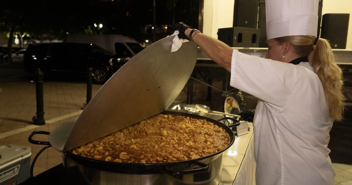 Un chef, vestido con uniforme blanco y un alto gorro de cocina, levanta la tapa de un recipiente grande para cocinar al aire libre, el cual contiene un plato a base de arroz, durante un evento comunitario nocturno.