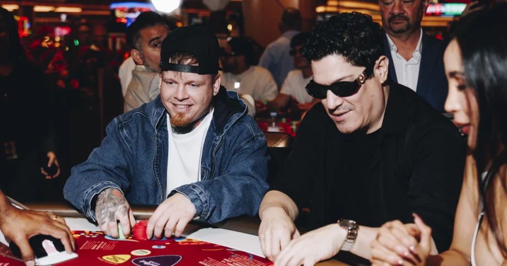 Un hombre con una gorra negra puesta al revés y una chaqueta vaquera, y otro vestido de negro y con gafas de sol, juegan a un juego de mesa de casino en el Seminole Hard Rock Hollywood, rodeados de otros clientes en el animado ambiente del casino.