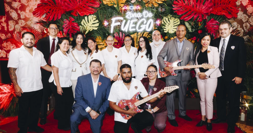A group of Seminole Hard Rock Hollywood team members and executives pose for a group photo in front of a colorful tropical display featuring a neon &ldquo;Zona de Fuego&rdquo; sign. Several team members hold custom-designed electric guitars decorated with vibrant artwork.