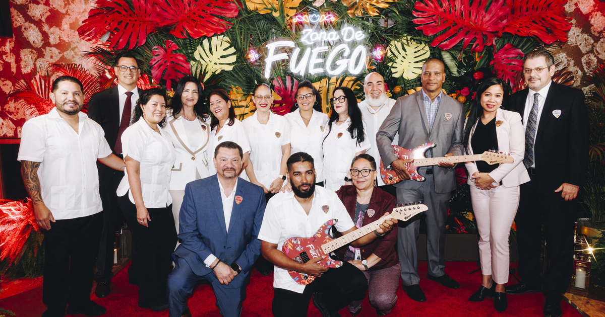 A group of Seminole Hard Rock Hollywood team members and executives pose for a group photo in front of a colorful tropical display featuring a neon &ldquo;Zona de Fuego&rdquo; sign. Several team members hold custom-designed electric guitars decorated with vibrant artwork.