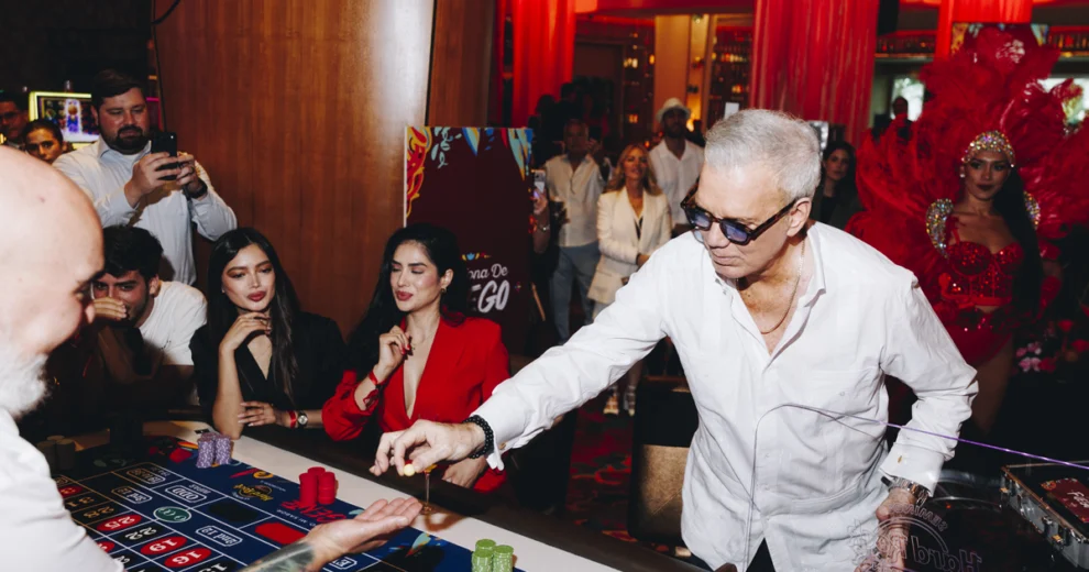 Un hombre con camisa blanca y gafas oscuras coloca una ficha en una mesa de ruleta del Seminole Hard Rock Hollywood, mientras dos mujeres observan desde el lado opuesto. Los clientes se reúnen alrededor de la mesa y, al fondo, se encuentra un artista con un traje rojo de carnaval y un elaborado tocado de plumas.