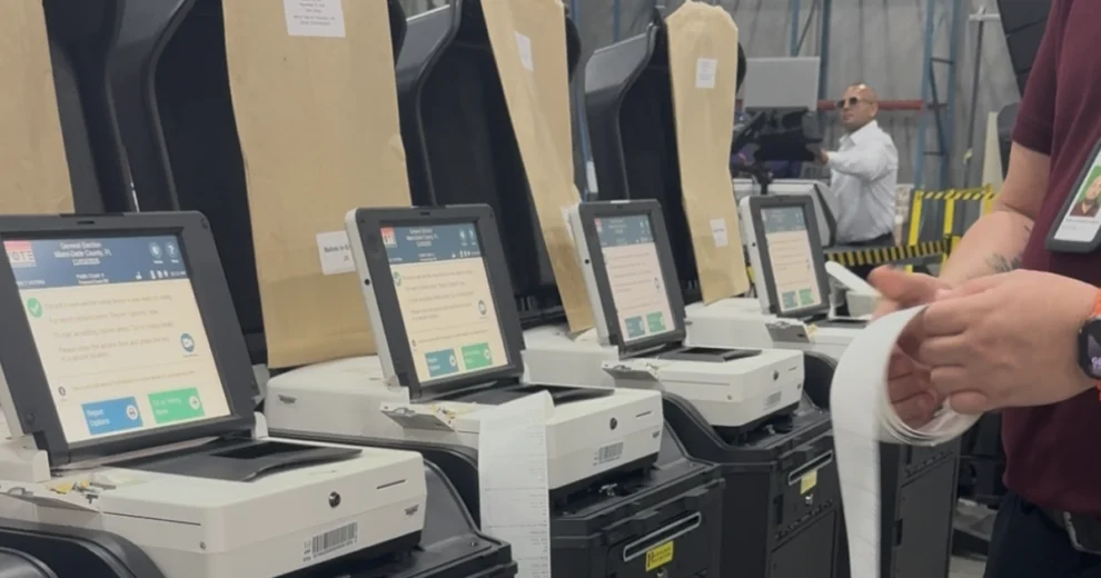 Una hilera de máquinas de votación electrónica, equipadas con pantallas táctiles e impresoras de papeletas, se encuentra dispuesta dentro de un almacén mientras el personal electoral prepara e inspecciona los equipos para las operaciones de votación.