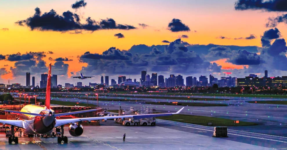 An airplane sits on the runway at Miami International Airport at sunset, with airport lights and the Miami skyline visible in the background beneath colorful clouds.