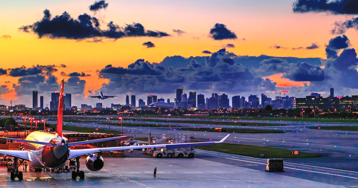 An airplane sits on the runway at Miami International Airport at sunset, with airport lights and the Miami skyline visible in the background beneath colorful clouds.