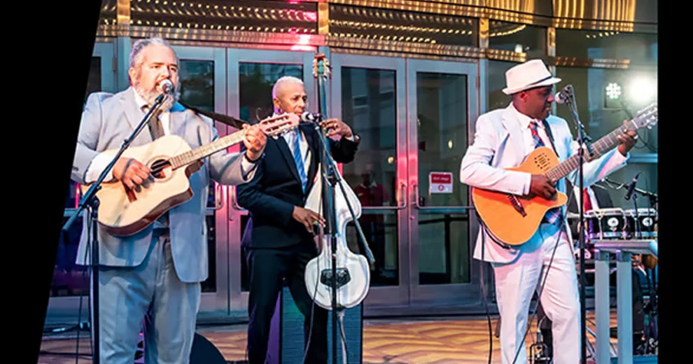 Unos músicos actúan en directo en Rhythm & Roots; tres guitarristas y cantantes tocan frente a la entrada de un edificio.