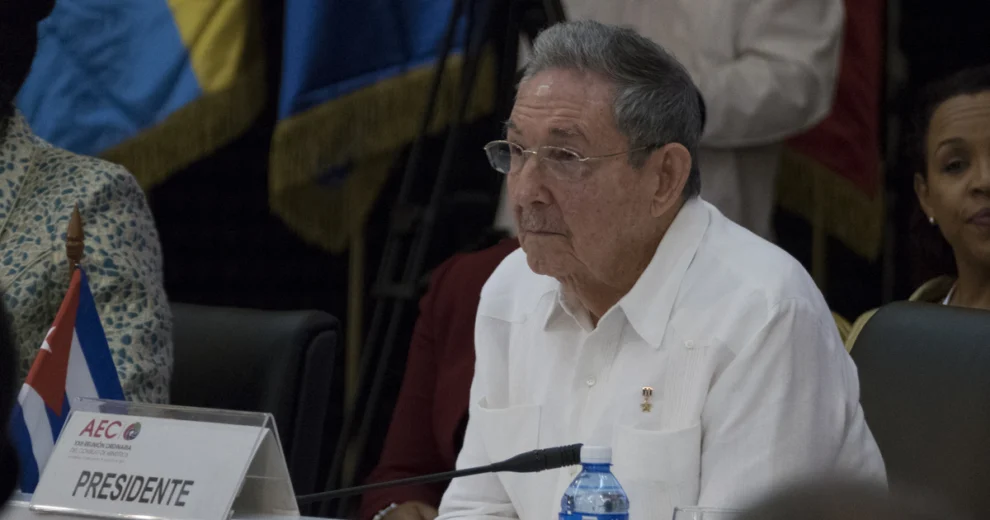 Raúl Castro sits at a conference table wearing a white shirt and glasses, with a Cuban flag beside him, a microphone, and a nameplate reading “Presidente.” Attendees and flags are visible in the background.