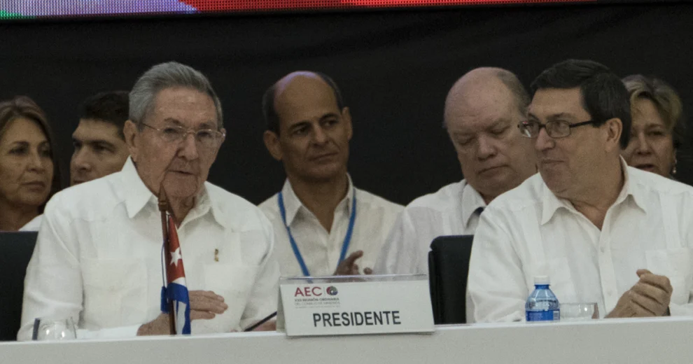 Raúl Castro está sentado junto a otros funcionarios en una conferencia; viste una camisa blanca y lleva gafas. Frente a él se encuentran una bandera cubana y una placa con la inscripción "Presidente", mientras que varios asistentes están sentados detrás de la mesa principal.