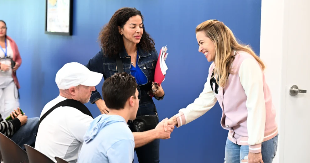 La comisionada del condado de Miami-Dade, Raquel Regalado, quien viste una chaqueta de color rosa y blanco, sonríe y estrecha la mano de un hombre sentado durante una visita a la comunidad. Cerca de ellos, una mujer sostiene unas carpetas, mientras otras personas permanecen sentadas al fondo.